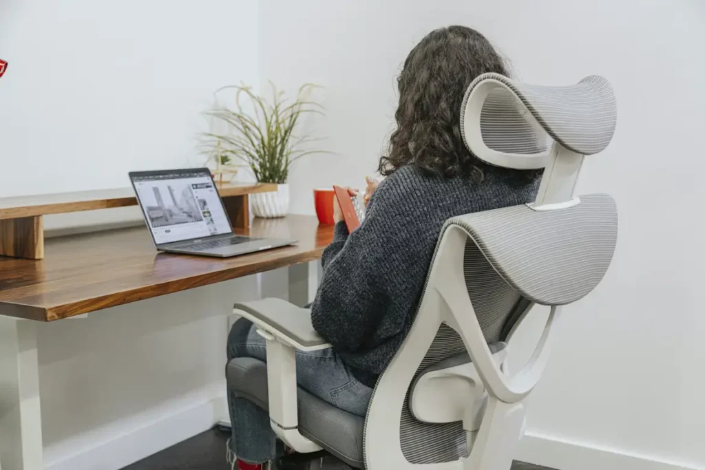A person with long, curly dark hair sits in a modern, ergonomic white and grey mesh office chair, facing a wooden desk. They are holding a small orange notebook. On the desk, there is a laptop, a potted plant, and a red mug.