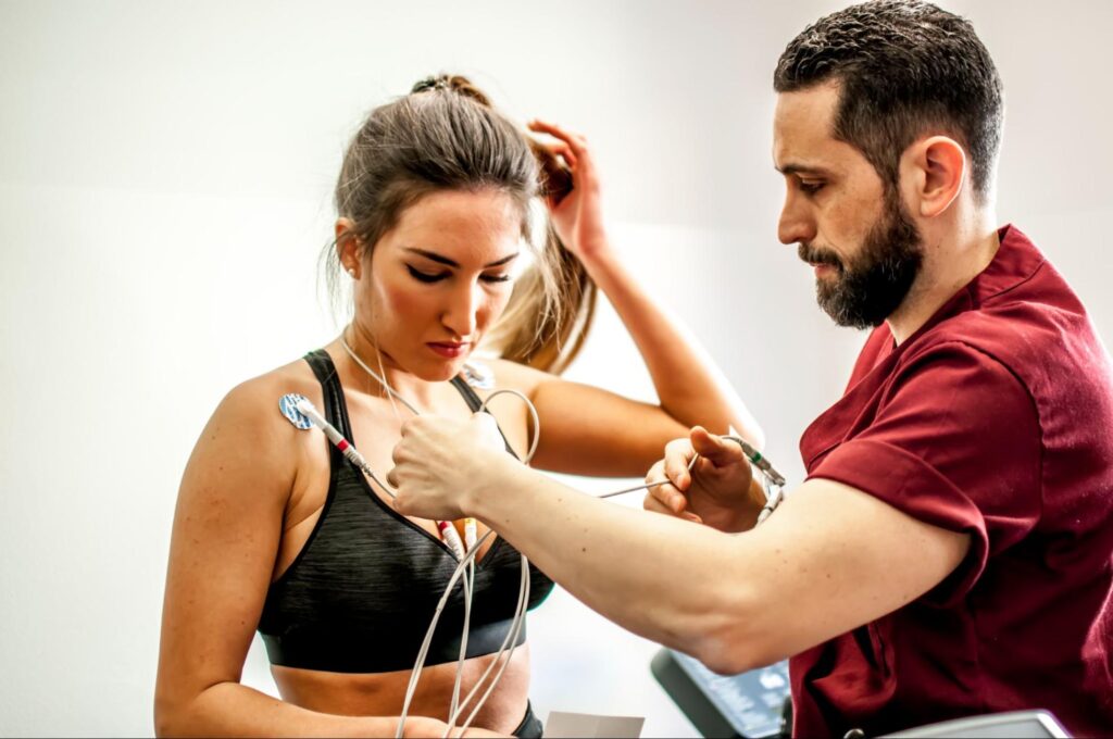 A healthcare provider in a red uniform places electrode pads and wires on a woman’s shoulders and chest as she prepares for an electric stimulation therapy session, wearing a black sports bra in a clinical setting.