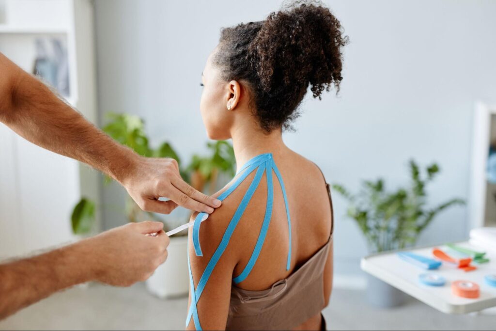 A clinician applies blue kinesiology tape to a woman’s upper back and shoulder, measuring and smoothing strips that fan out from the base of her neck toward her deltoid in a bright therapy room.