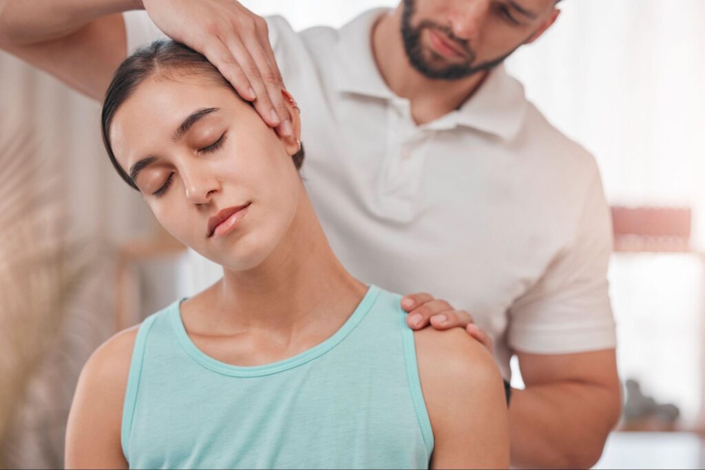 A chiropractor in a white polo shirt gently stretches a young woman’s neck to the side while she remains seated with eyes closed, receiving cervical traction therapy in a softly lit clinic environment.