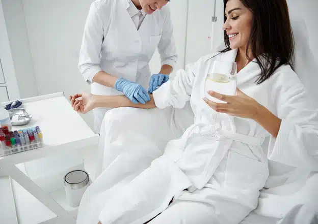 A woman in a white robe sits comfortably while a healthcare professional prepares her arm for an IV treatment, holding a glass of water and smiling.