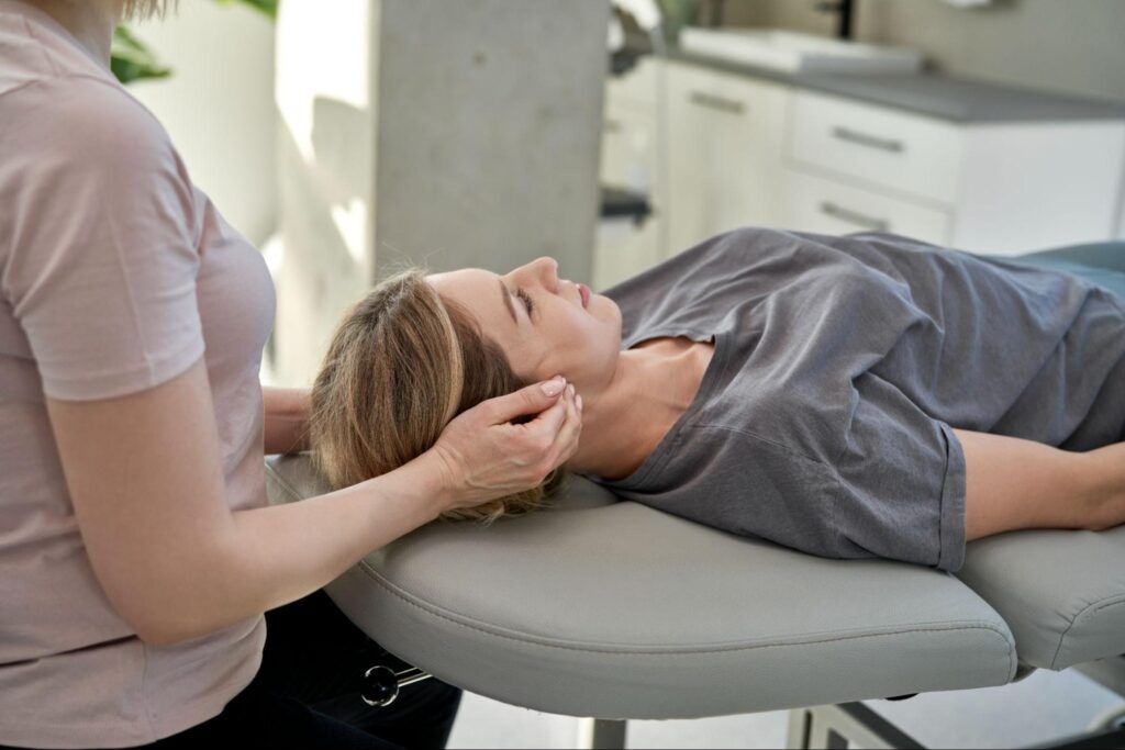 A therapist seated beside a treatment table gently cups and supports a woman’s head as she lies face-up during a medical massage session in a bright clinic room.