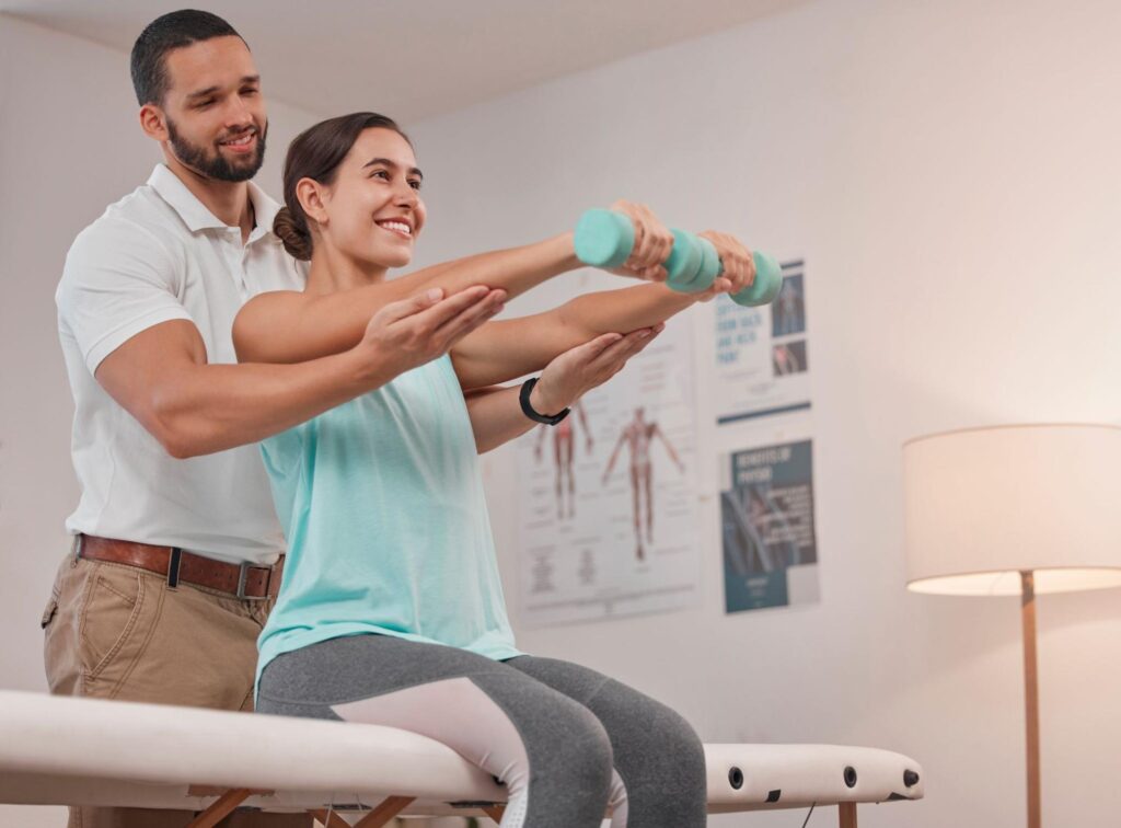 A male therapist stands behind a seated female patient, guiding her arms as she holds light teal dumbbells straight in front of her, performing a shoulder-strengthening exercise in a bright therapy room with anatomical posters and a floor lamp.