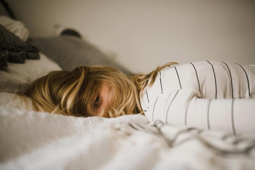 A woman with blonde hair lying face down on a bed, wearing a white shirt with black horizontal stripes. She looks tired or fatigued, with her face partially turned towards the camera and her hair slightly covering one eye. The bedding is light-colored and soft-focused in the foreground.
