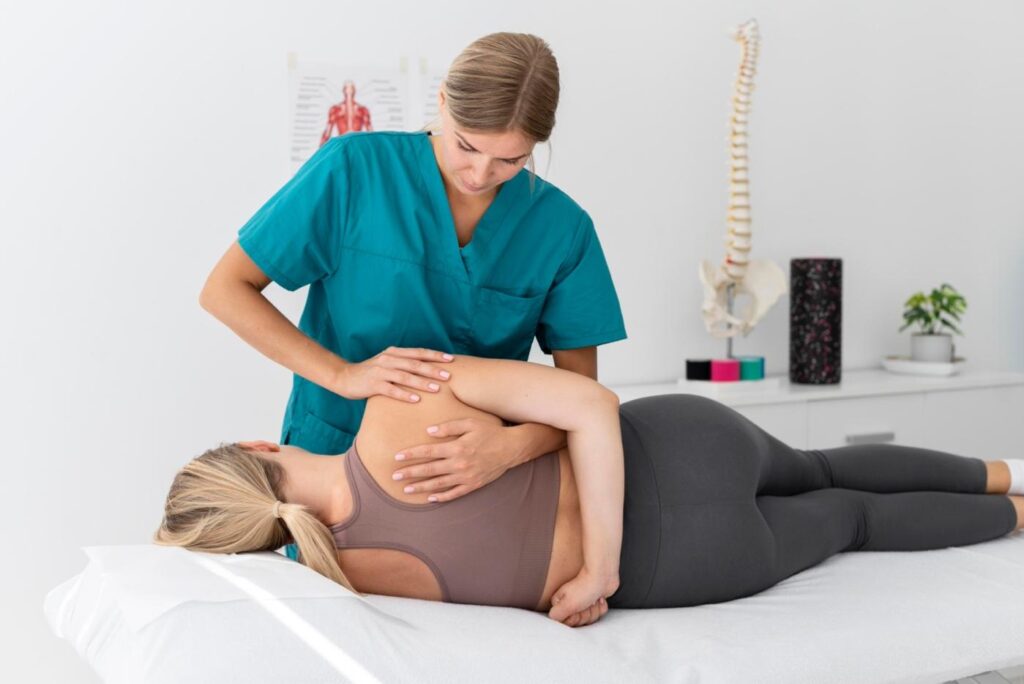 A physical therapist in teal scrubs applies a manual shoulder and upper-back mobilization technique to a female patient lying on her side on a treatment table, with a spine model and foam roller visible in the bright clinic background.
