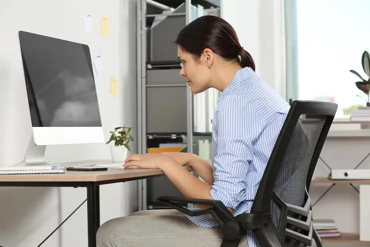 Woman sitting at a desk with poor posture while working on a computer, leaning forward in an office chair, illustrating the effects of prolonged sitting and spinal strain.