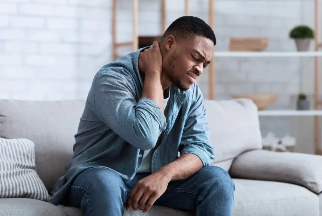 Man sitting on a couch holding his neck and wincing in pain, illustrating symptoms of whiplash
