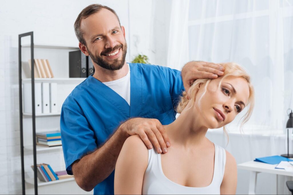 A smiling therapist in blue scrubs gently stretches a woman’s neck by holding her head and supporting her shoulder, demonstrating a non-invasive manual therapy technique in a bright treatment room.