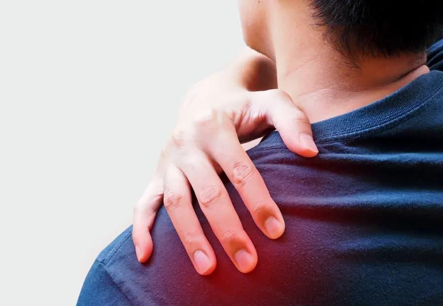 A person wearing a dark blue shirt is seen from behind and slightly turned to the side. They are using their right hand to hold their left shoulder, indicating pain or discomfort. The area around the left shoulder is highlighted with a red glow to emphasize inflammation or pain. The background is a neutral light grey.