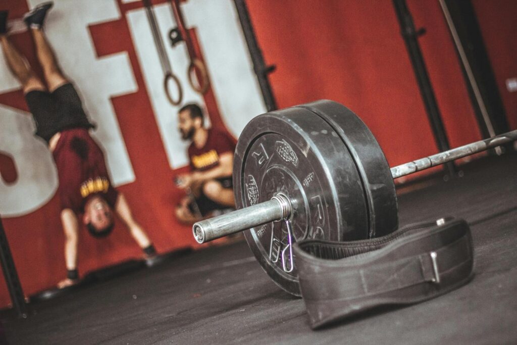A loaded barbell with black weight plates and a lifting belt resting on the gym floor, with a blurred background showing a coach observing an athlete performing a handstand push-up against a red and white wall decorated with gym rings.