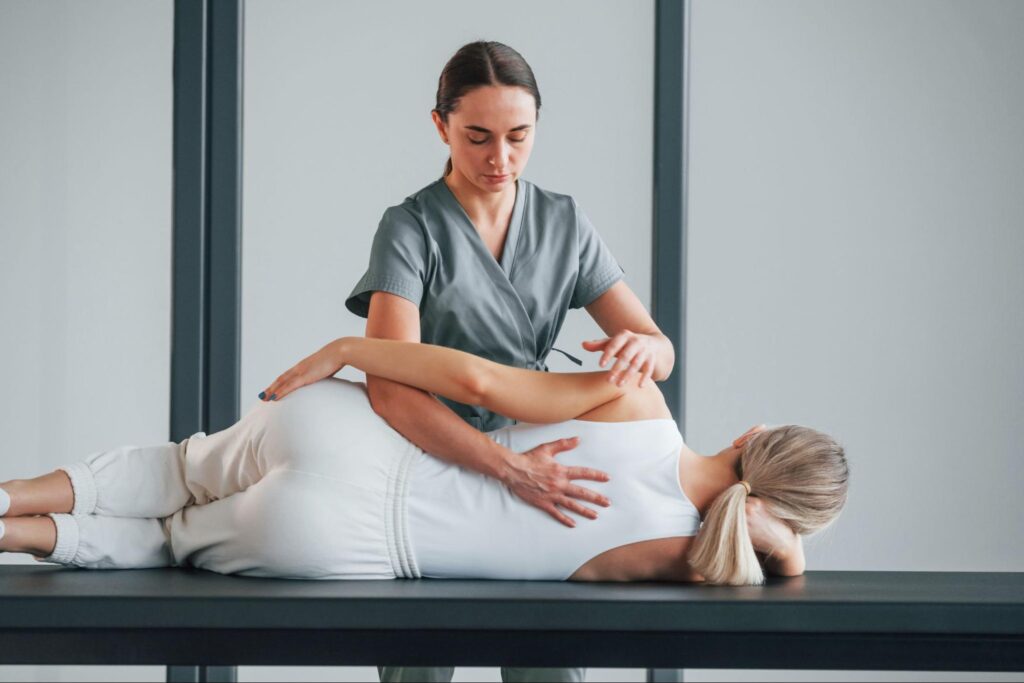 A physical therapist in gray scrubs applies targeted trigger-point pressure to a client’s upper back while she lies on her side on a treatment table, using hands to palpate and release muscle tension. 2/2