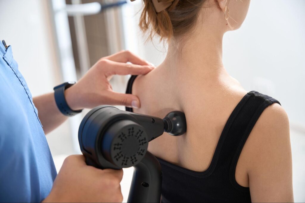 A therapist in blue scrubs holds a handheld ultrasound therapy device against the upper back of a seated patient wearing a black tank top, gently positioning the treatment head on her shoulder blade area.
