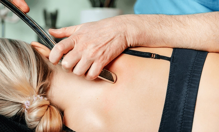 Close-up of a practitioner’s hands using a stainless steel Graston instrument to apply soft-tissue mobilization along the upper back near a patient’s shoulder blade, who is lying face-down on a treatment table.