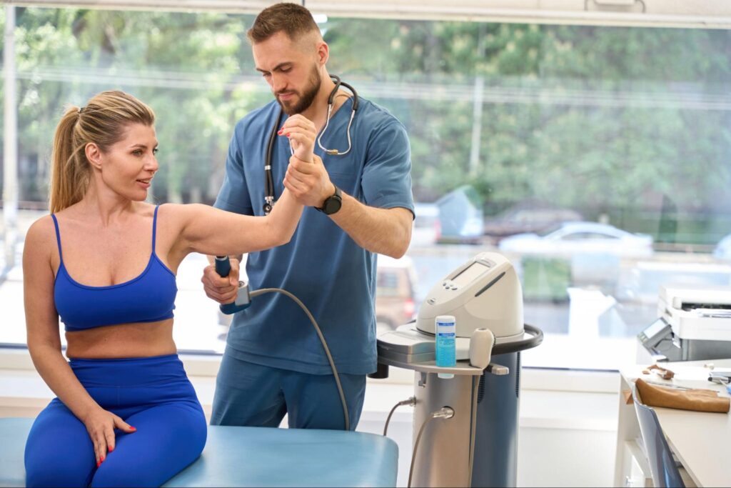 A clinician in blue scrubs uses a handheld therapeutic ultrasound device on a seated woman’s raised arm, guiding the ultrasound head over her forearm muscles in a sunlit clinic with the ultrasound machine beside them.