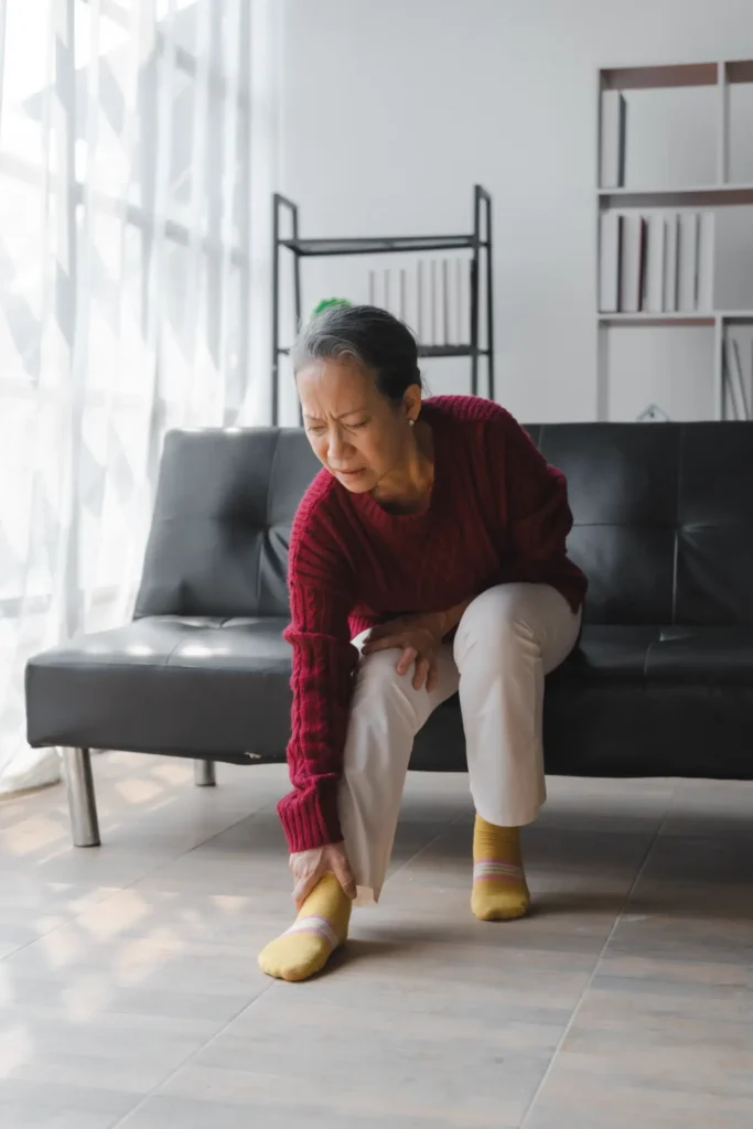 Elderly woman holding ankle in pain, seated in a modern clinic, Manhattan NYC