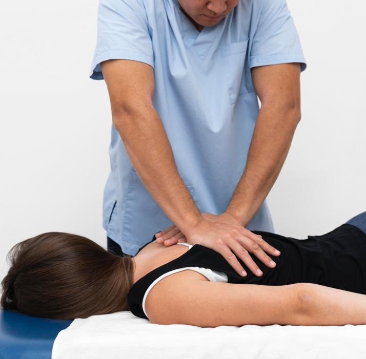 Photo of a man in a light blue medical scrub top performing a back adjustment on a woman lying face down on a therapy table. The man uses both hands to apply pressure on the woman’s upper back. The woman has brown hair and is wearing a black sleeveless top. The background is plain white. The image depicts a chiropractic or manual therapy session focused on proprioception and musculoskeletal adjustment.