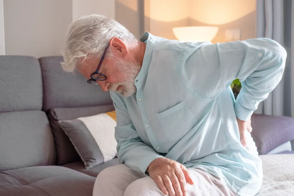 Elderly man with back pain, potential chiropractic patient, sitting on a sofa in a Manhattan clinic setting