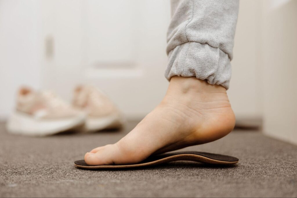 Close-up of a bare foot pressing down onto a brown, contoured orthotic insole on a carpeted floor, with a pair of sneakers blurred in the background.