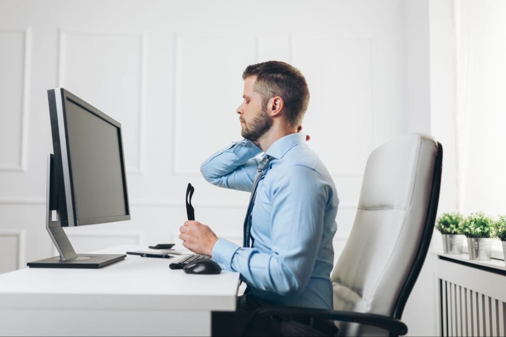 A man in a light blue dress shirt sits at a desk with a computer, holding his neck in discomfort with his eyes closed, suggesting neck strain or poor posture from prolonged screen use.