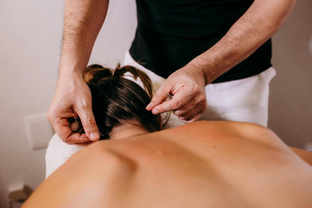 Close-up of a practitioner’s hands inserting fine acupuncture needles into the back of a patient’s neck, with the patient lying face-down on a treatment table.