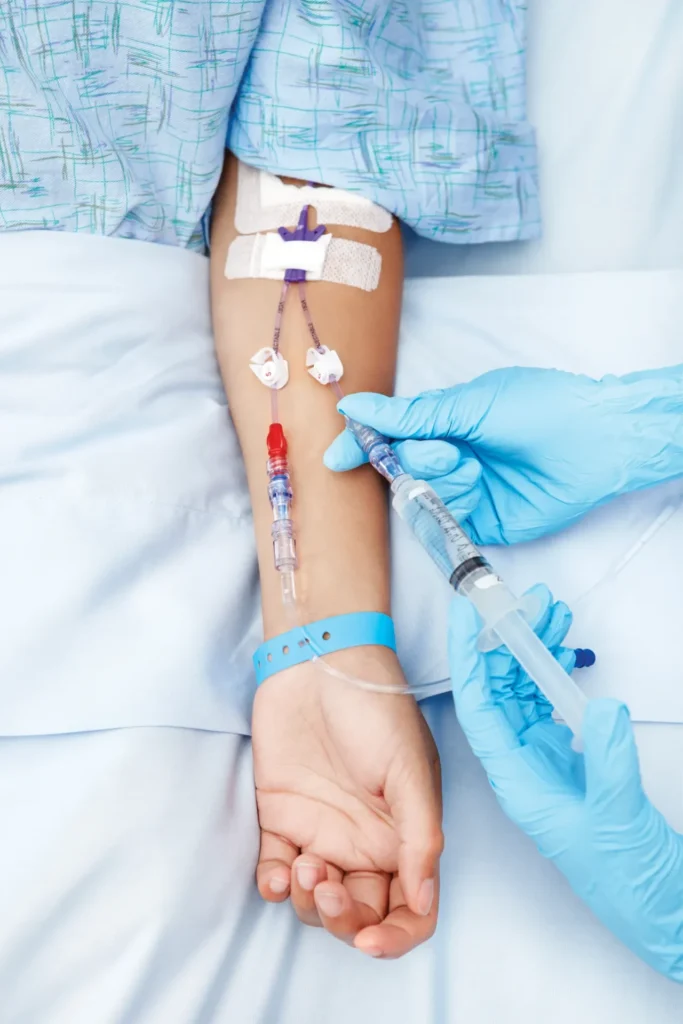 A healthcare professional wearing blue gloves administers medication through an IV line inserted in a patient’s arm in a hospital setting.