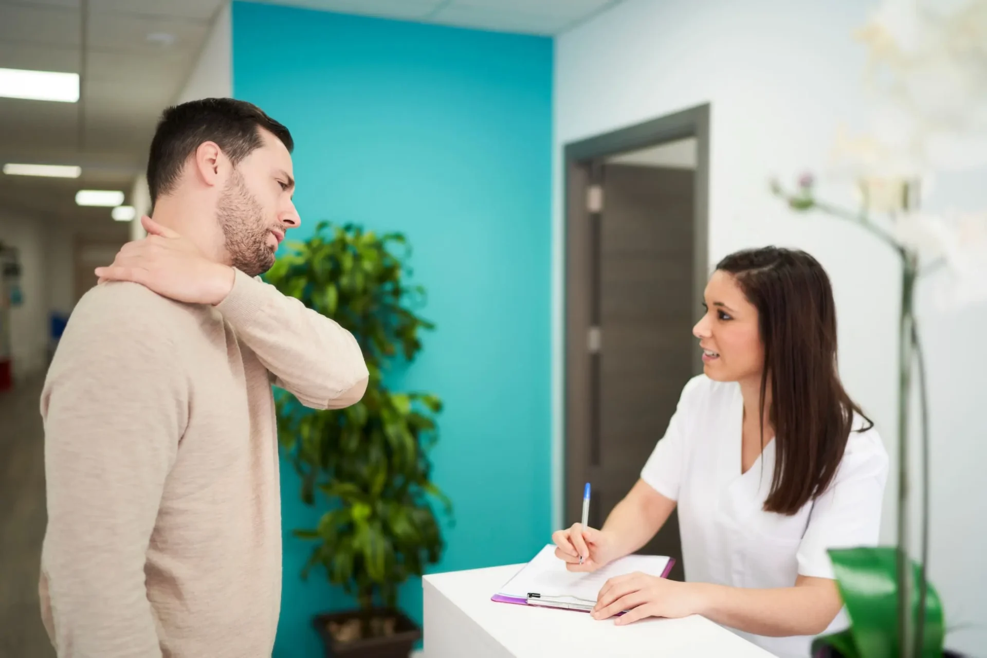 Man checking in at a clinic reception desk with a female nurse or assistant, pointing to his painful neck/shoulder.