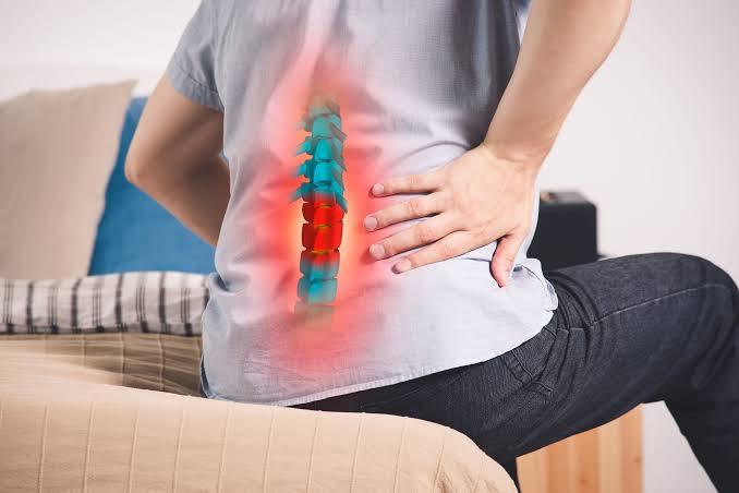 A person sitting on the edge of a bed, holding their lower back with one hand in discomfort. The image shows a digitally superimposed illustration of the lumbar spine, highlighting some vertebrae in red to indicate pain or spinal degeneration. The background includes a beige quilt and pillows on the bed, suggesting a home setting. The image conveys symptoms or awareness of lower back spinal degeneration or related pain.