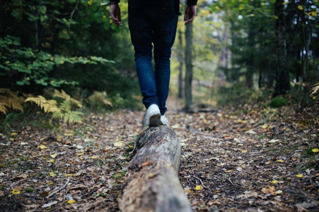 Photo of a person walking on a fallen tree trunk in a forest. The person is wearing dark jeans and white sneakers, and the focus is on their feet balancing on the narrow trunk. The surrounding area has dirt, leaves, and green trees, indicating an outdoor natural setting. The image conveys balance, coordination, and proprioception in a natural environment.
