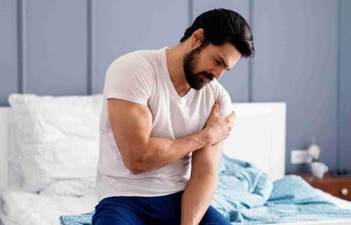 A man sitting on a bed, clutching his right shoulder with his left hand and showing a grimace of pain on his face. He is wearing a white t-shirt and blue pants. The image conveys discomfort or pain in the shoulder area, potentially related to a clavicle fracture or shoulder injury.