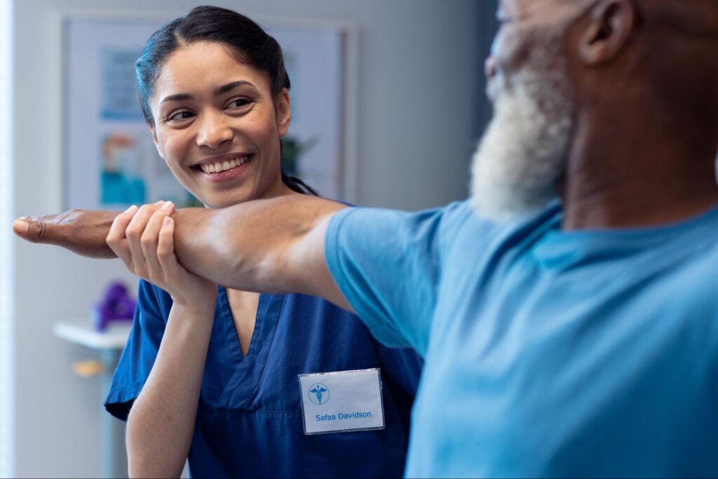 A smiling therapist, wearing blue scrubs and a name tag, gently supports and stretches an older man’s extended arm in a clinical treatment room.