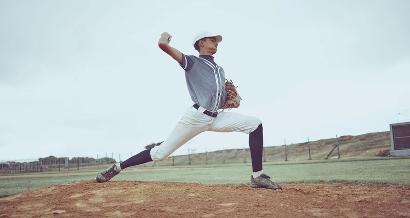 Baseball pitcher in white uniform n mid-pitch delivery, demonstrating full extension of arm during throwing motion - a movement that can stress the elbow and ulnar nerve, relevant to cubital tunnel syndrome.