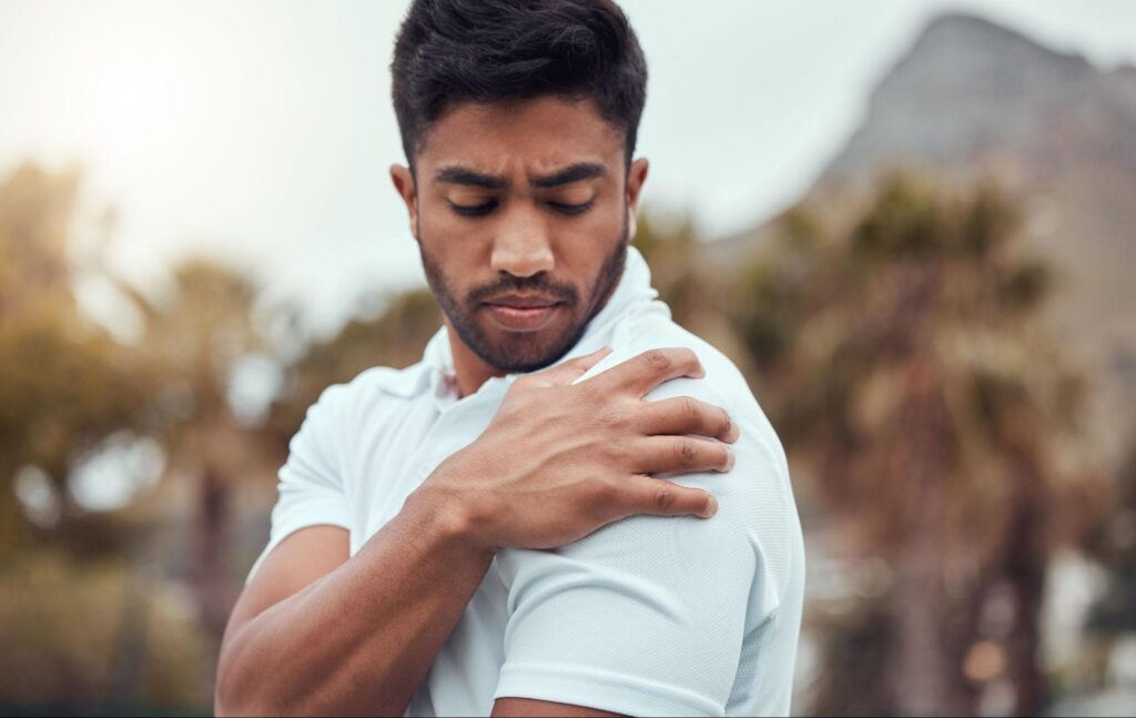 A man outdoors wearing a white polo shirt holding his right shoulder with a pained expression, indicating shoulder discomfort or injury.