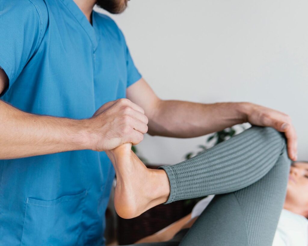 Photo of a physiotherapist or medical professional in blue scrubs stretching a patient's leg and ankle. The patient is lying down on a treatment bed, and the professional holds the patient's ankle with one hand and supports the knee with the other, bending the leg gently. The focus is on the hands and the leg stretch, showing a typical physical therapy or rehabilitation session for knee pain or mobility improvement. The setting appears to be a clinical or therapy room with plants blurred in the background.
