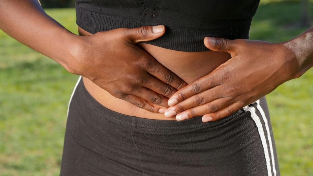Close-up image of a person outdoors, wearing black sportswear, holding their lower abdomen with both hands. The background shows green grass. The image suggests symptoms or conditions related to nausea or abdominal discomfort.