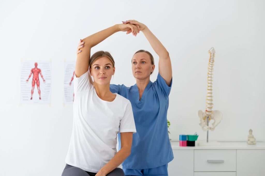 A physical therapist in blue scrubs assists a seated woman in a white t-shirt with an over-head arm stretch, holding her elbow and hand for support in a bright clinic room with an anatomical chart and a spine model in the background. 3/3