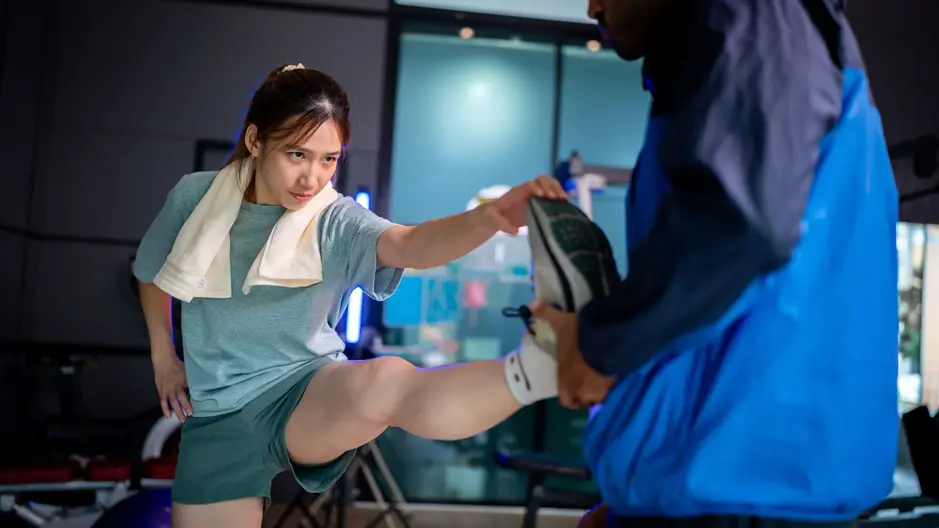 Female runner in gym doing dynamic leg stretch with coach support during NYC Marathon taper week — injury prevention warm-up.